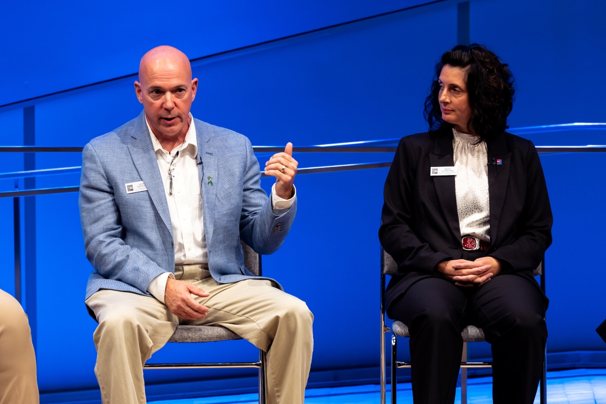 A bald man in a blue blazer holds up his left hand and speaks. A woman beside him in a black suit and white shirt, hands folded, listens.