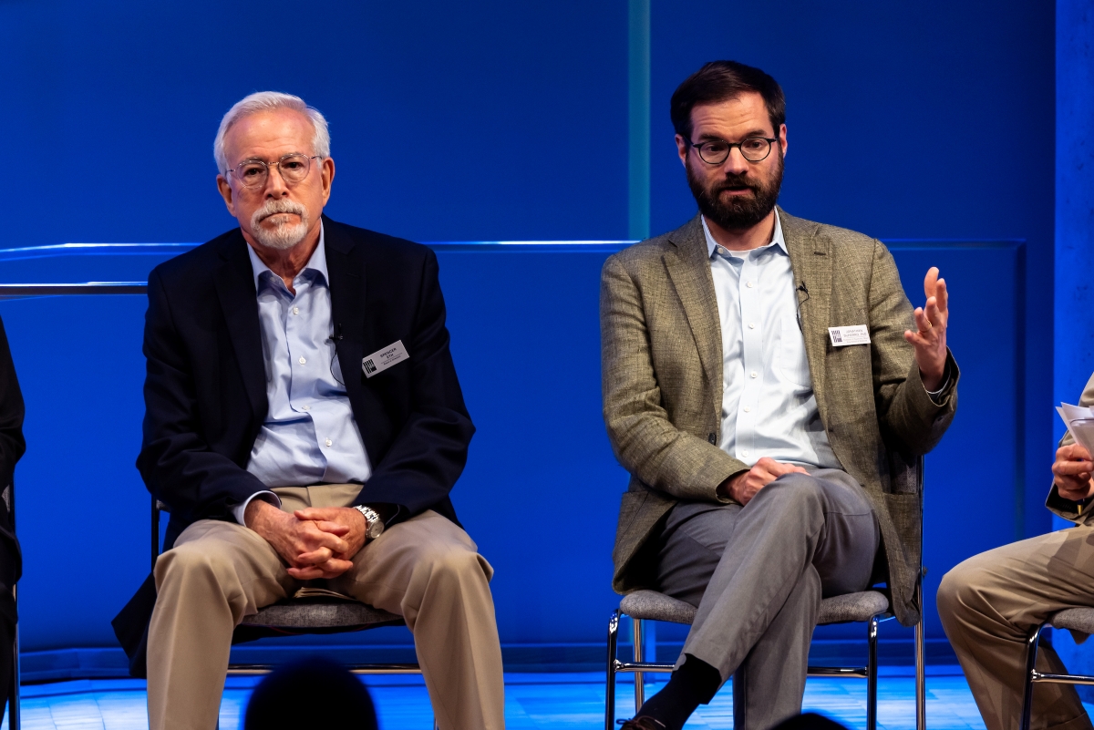 Two men sit on stage. The man on the left  faces the camera, not speaking, hands folded, while the man on the right gestures and speaks.