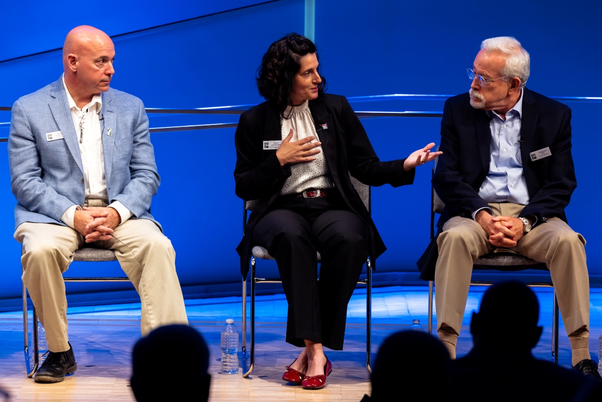 Three people sit on stage. The woman in the middle speaks and gestures while two men listen on either side.