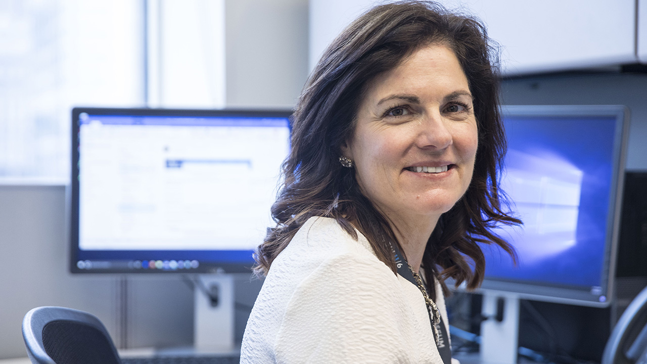 A female volunteer with brown hair looks towards the photographer with a smile. Two computer screens are on behind her. 