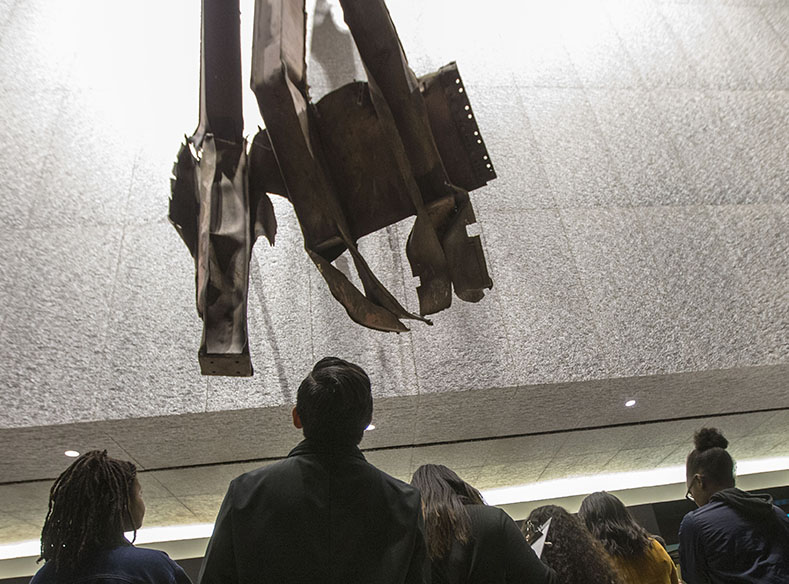 Six dark-haired men and women looking up to an artifact of a damaged 3-piece steel beams mounted on textured aluminum claddings. 