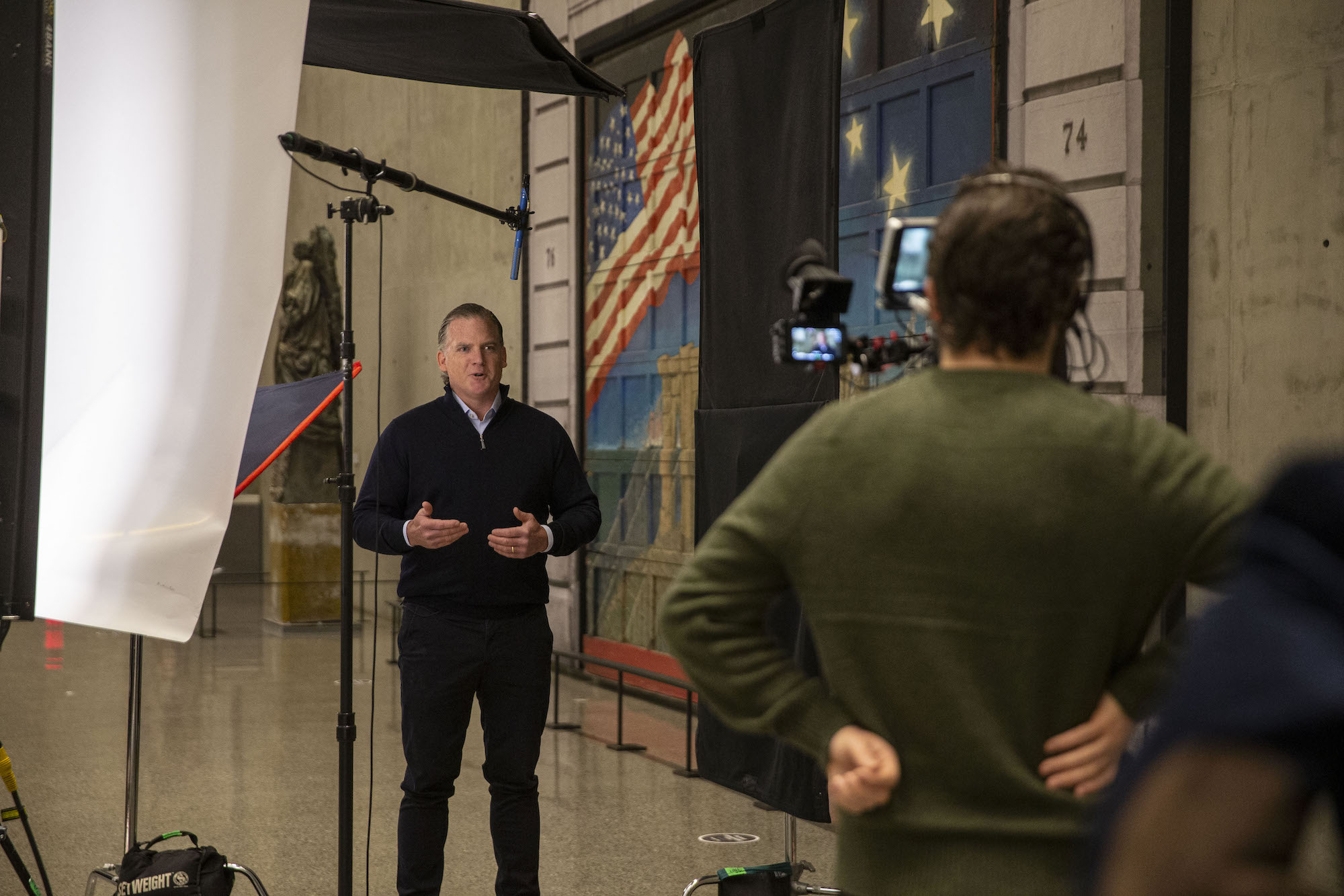 A man in a dark sweater and pants speaking to camera man 