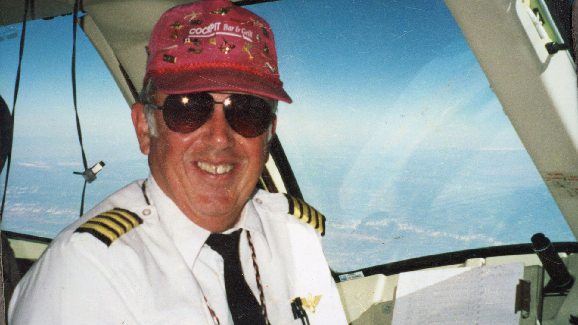 A smiling man in a cockpit, wearing a pilot's uniform and a red baseball cap 