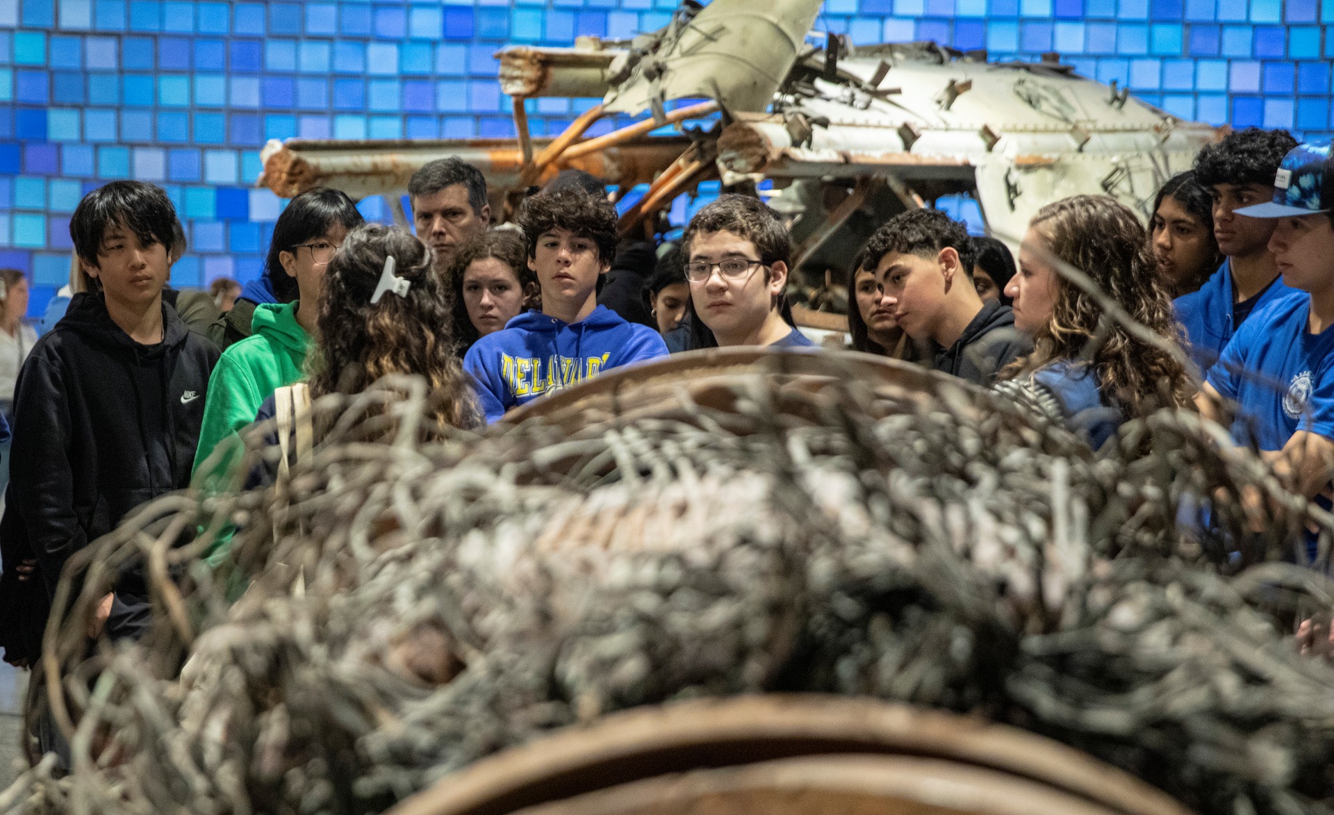 a group of students stands behind a large metallic artifact with the blue Spencer Finch installation visible behind them