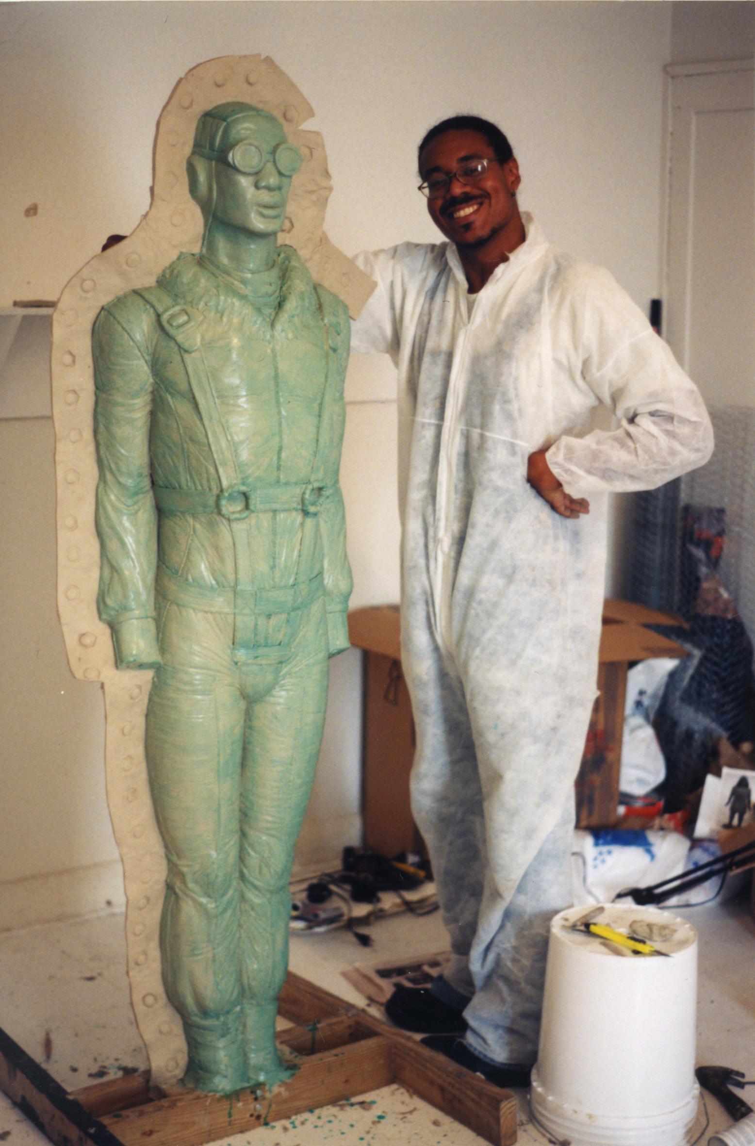 A smiling man in white coveralls stands with his arm around a cast of his own body, dressed like a WWII pilot