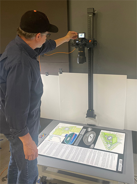 A man in a baseball cap, wearing jeans and a dark untucked shirt, adjusts a light over design boards that show proposals for the memorial