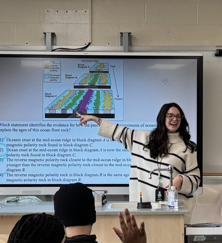 A teacher stands in front of her students, pointing to a presentation on screen