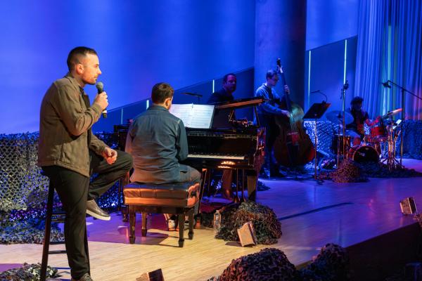 A person speaks into a microphone, the backs of others are visible on stage, including a pianist