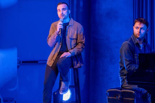 a man sits on a stool and speaks into a mic. he wears a brown jacket and blue pants. the piano player is slightly visible to the right, facing the man from a rear angle