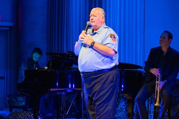 the same man from the previous slide, but a bit farther away. The man speaks into the microphone, holding it with two hands.