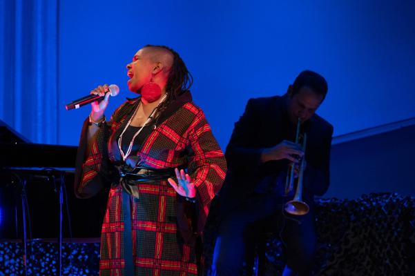 The woman in the blue and red square pattern dress sings with her right hand holding a microphone; there is a blue background and someone as a shadow is visible behind her