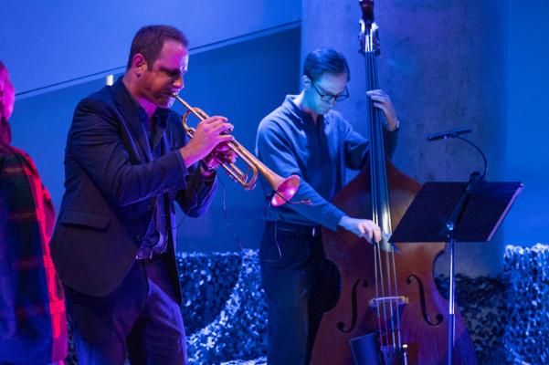 The trumpet player and an upright bass player are visible with a music stand in the foreground. The stage is blue and darkly lit.