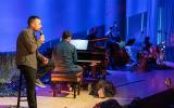 A person speaks into a microphone, the backs of others are visible on stage, including a pianist