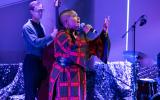 A woman in a blue and red dress raises her arms and sings