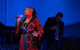 The woman in the blue and red square pattern dress sings with her right hand holding a microphone; there is a blue background and someone as a shadow is visible behind her