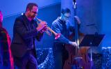 The trumpet player and an upright bass player are visible with a music stand in the foreground. The stage is blue and darkly lit.