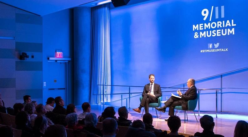 Two men take part in a discussion at the Museum. The auditorium is lit blue and spotlight is on the two men as they sit on stage. Silhouetted audience members watch from the left.