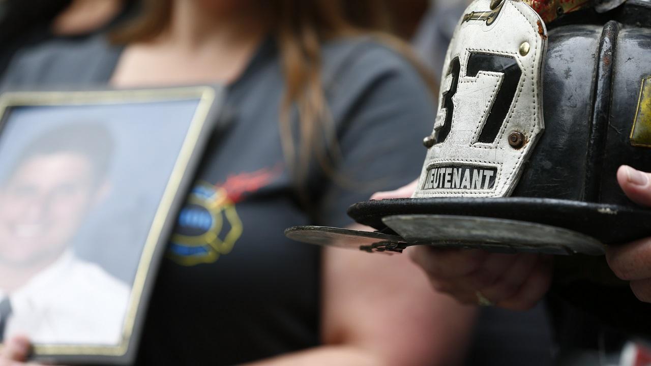 In focus to the right are two hands holding a firefighter helmet with the number 37 and the word lieutenant on it. Out of focus to the left is a woman holding a framed photo of a man.