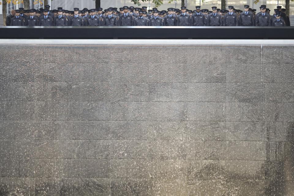 Dozens of probationary firefighters stand beside a reflecting pool on the 9/11 Memorial plaza. Water cascades down to the pool below them.