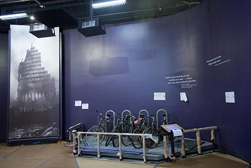 A bicycle rack from Vesey Street is displayed in the Museum before its opening. The rack was damaged and covered in dust and debris during the 9/11 attacks. A large image from Ground Zero stands to the left of the rack.