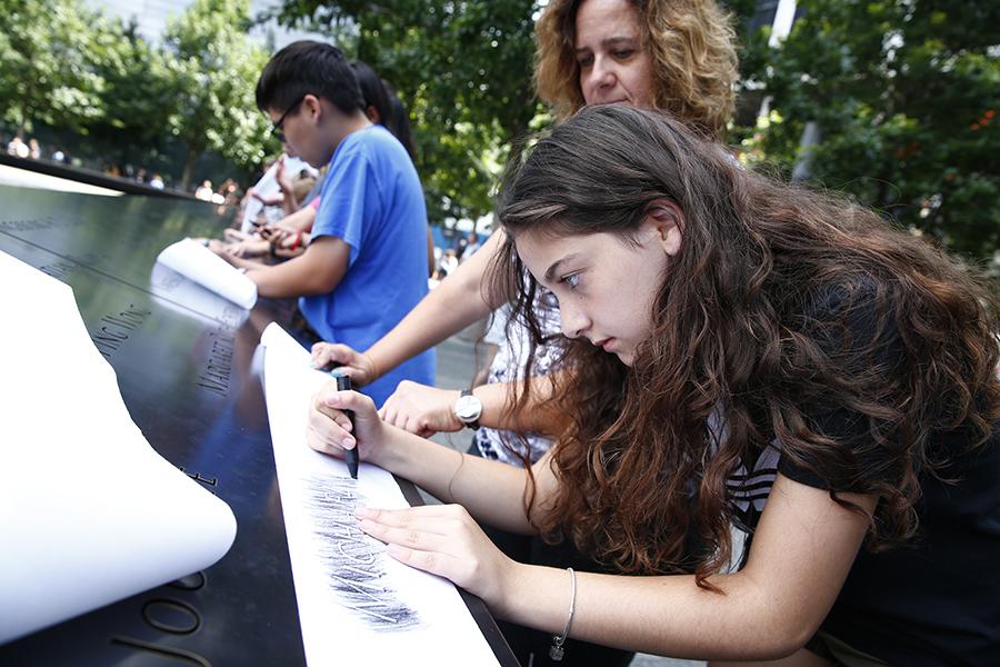 A girl stands next to a woman as she makes a charcoal rubbing at a name etched on a bronze parapet at the Memorial. Other young visitors do the same behind her.