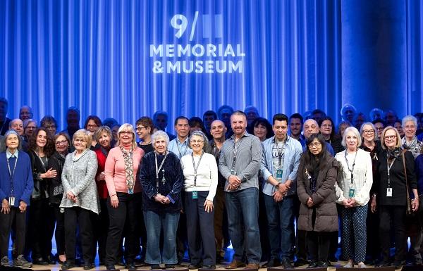 Dozens of volunteers, primarily older adults, are seen onstage at the Museum auditorium during the National Volunteer Week ceremony and reception. A blue curtain behind them has the 9/11 Memorial & Museum logo projected on it.