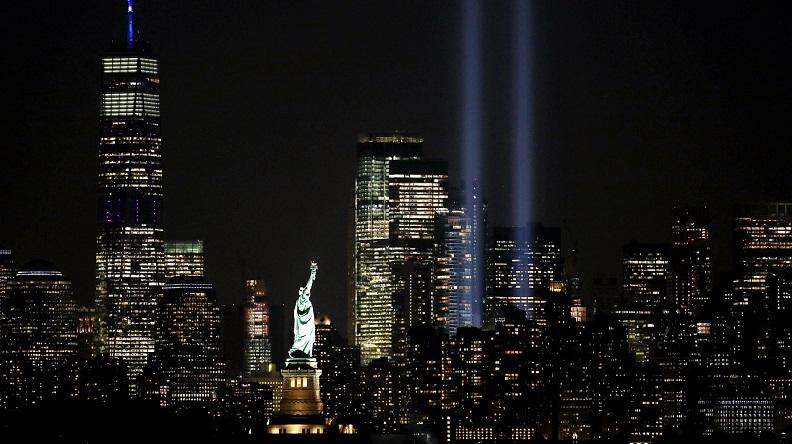 In this photo, the twin beams of the Tribute in Light installation shine skyward at night with the Statue of Liberty in the foreground and One World Trade visible to the right of the beams.