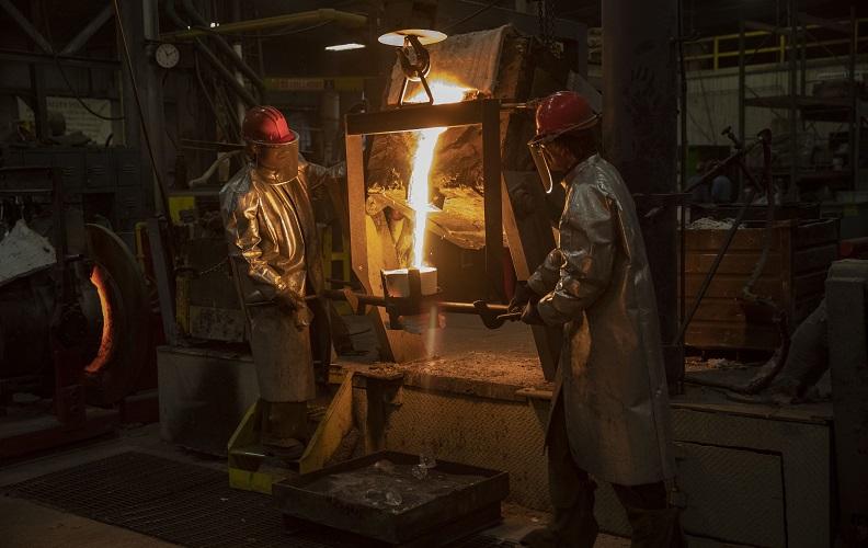 In this photo, two metal workers melt 9/11 World Trade Center steel into hot liquid at a foundry while wearing red hardhats and metallic coverings.