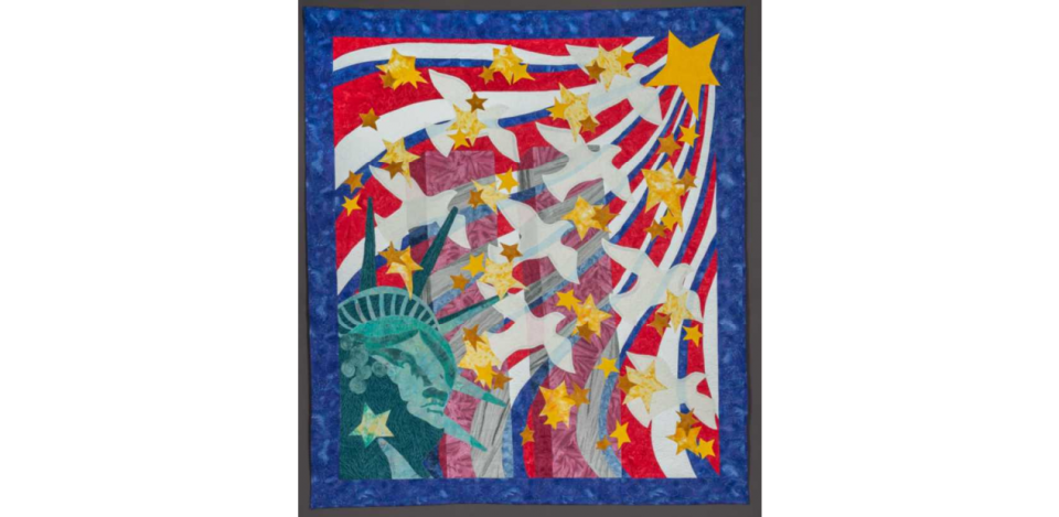 Handmade textile quilt depicts the Twin Towers at center, ghostlike in their transparency. The crowned head of the Statue of Liberty is represented in the lower left corner of the quilt, and from it white doves and yellow stars emanate. These images are set against waves of red, white, and blue.