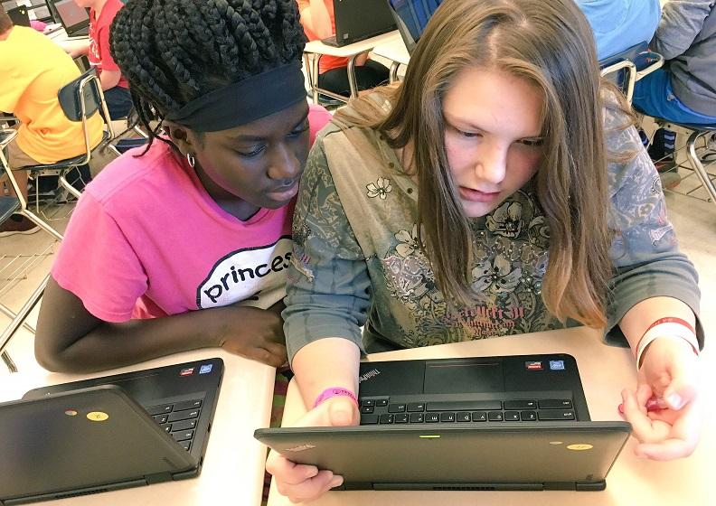 Two young girls sit in a classroom and look at a lesson on a laptop screen.