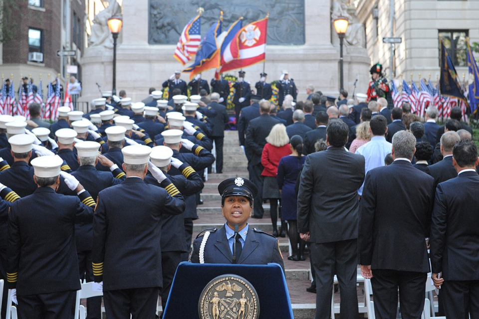 A woman in a navy blue firefighter's uniform speaks at a podium. Behind her, the backs of program attendees saluting six uniformed men on a stage.