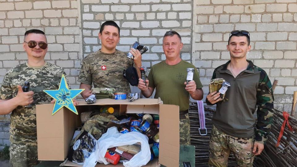 Four men with cropped dark hair, dressed in military fatigues, one holding a blue and yellow hand-painted wood star, stand against a white brick wall giving "thumbs up." An open box of canned food and supplies is visible in front of them. 